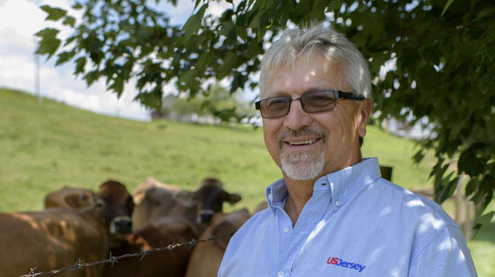 A man wearing a light blue shirt with “US Jersey” above the chest pocket stands smiling in front of several cows.