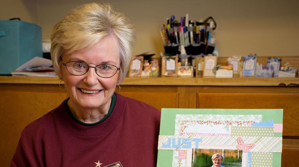 An elderly woman holds a scrapbook page while sitting behind a table full of several more pages.