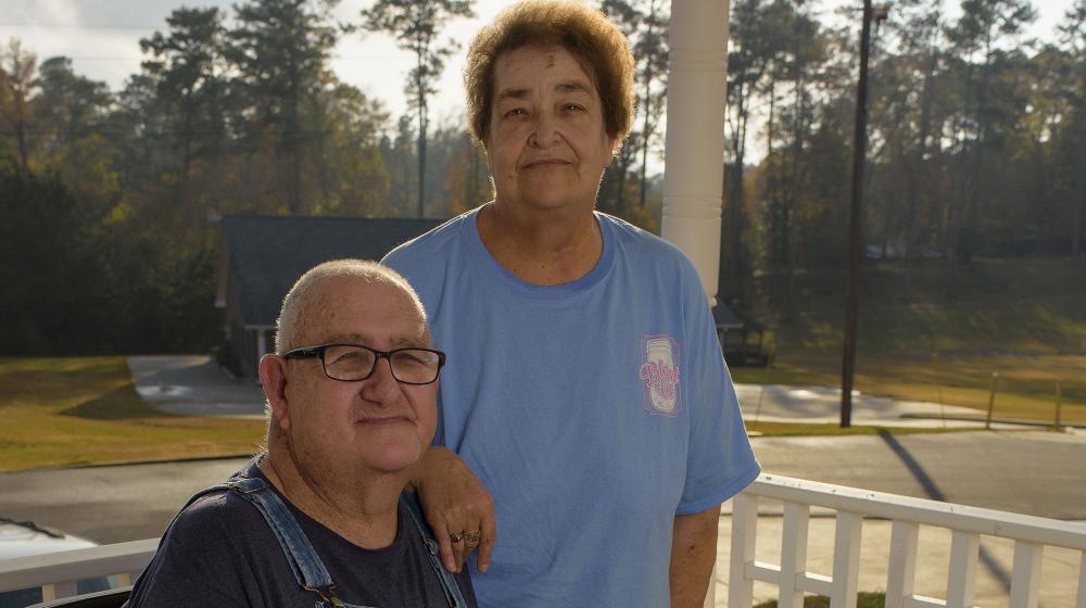 A gray-haired man wearing overalls and glasses sits beside a raised salad table, and a black-haired woman stands behind him, wearing a blue T-shirt, with her hand on his shoulder.