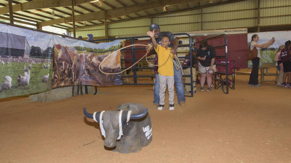 Amite County 2nd grader Elijah Johnson and Extension Agent Lionel Brown, Jr. A young boy throws a rope toward a fake bull with the help of an older man standing behind him.