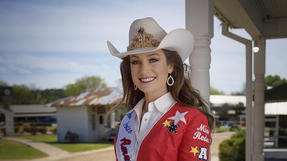 A young woman wearing a white cowboy hat and a red embroidered blazer stands on a wooden deck.