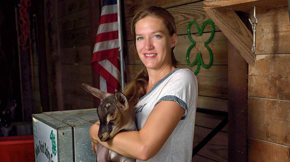 A blonde woman in a white shirt and ponytail stands in front of a wooden wall and holds a brown, black, and white baby goat. 