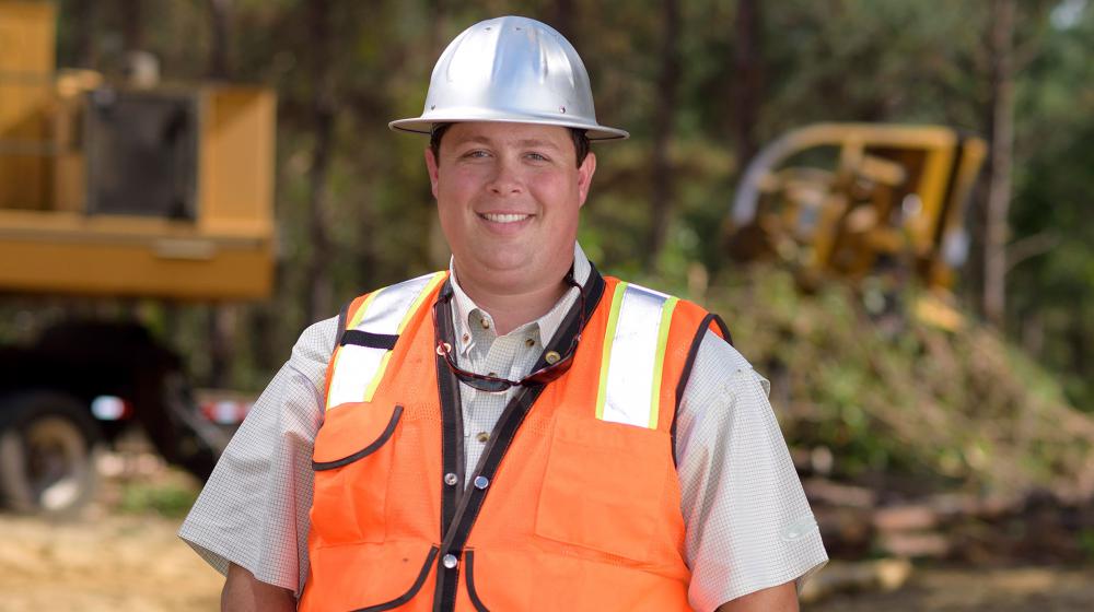 A young man with a metal construction hat and bright orange vest stands in front of his work site with his hands tucked into his blue jeans.