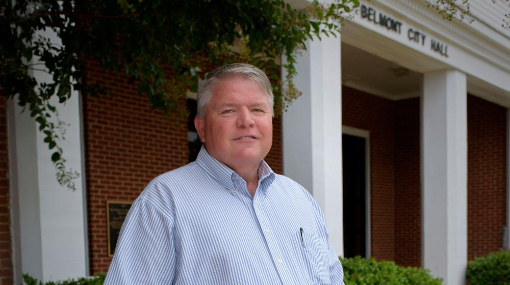 A man wearing a light blue and white striped shirt and blue jeans stands in front of Belmont City Hall.