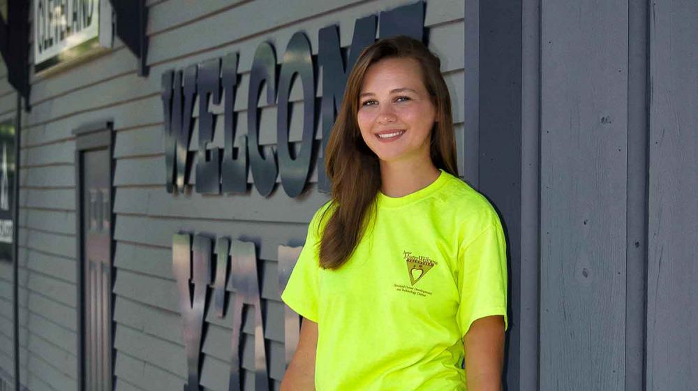 A teen with brown hair and wearing a green Junior Master Wellness Volunteer T-shirt stands in front of the Cleveland, Mississippi, Welcome Center.