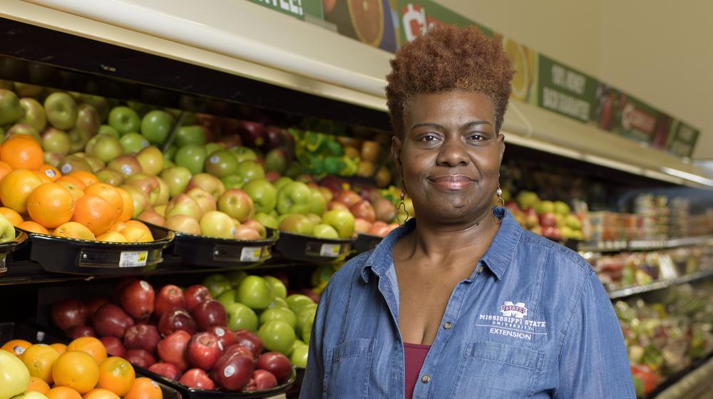 A woman wearing a blue collared jacket stands in the fruit aisle of a grocery store.