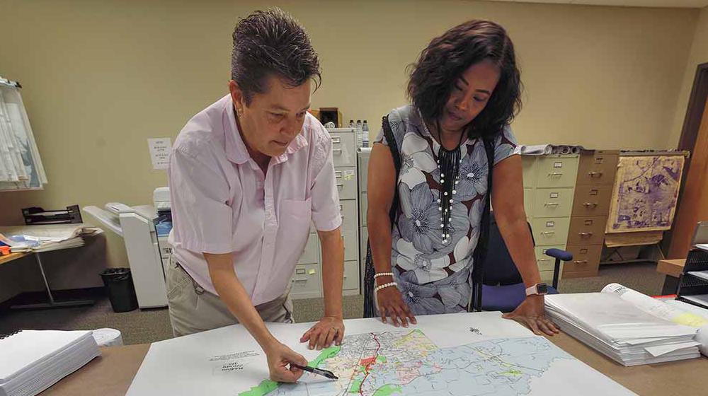 On the left, a woman with short hair and a light pink, collared shirt leans on the table, holding a pen above an area of a map. On the right another woman wearing a blue floral dress leans on the table and looks at it.