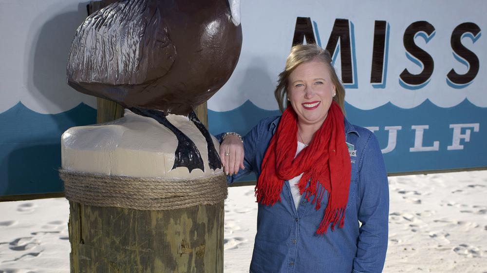A smiling woman with blonde hair, a red scarf, a denim shirt, and black pants rests her arm on the side of a “Welcome to the Mississippi Gulf” sign.