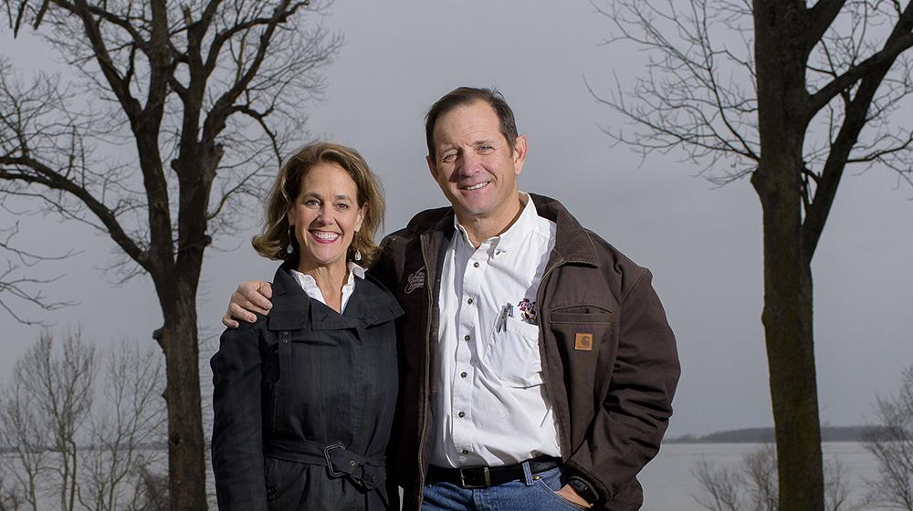 A man stands next to a woman with his arm around her shoulders while they both smile at the camera; a lake rests in the background.