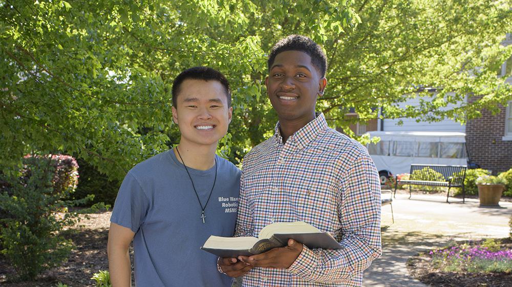 A boy wearing an orange and blue plaid shirt and holding an open book stands next to another boy wearing a blue shirt.  Photo credit: Kevin Hudson