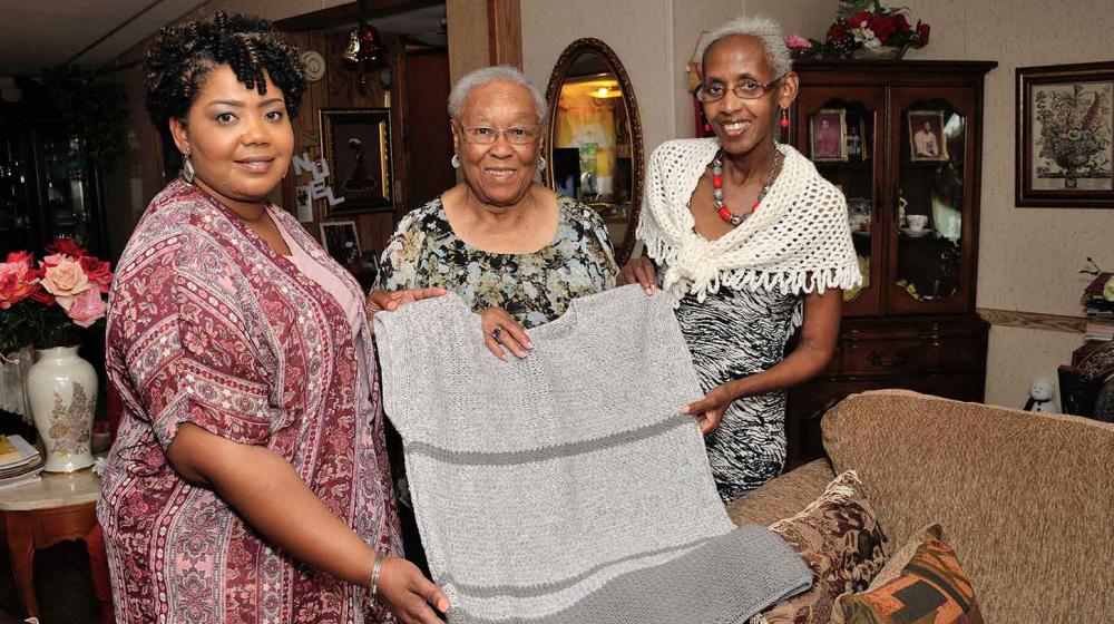 : On the left, a younger woman wearing a patterned pink shirt holds one side of a crocheted grey shirt shirt. In the middle, and older woman smiles. On the right, another older woman with grey hair and a crocheted piece of clothing smiles and holds the shirt. 