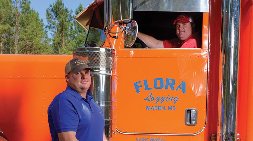 A man in a blue shirt stands in front of an orange semi-truck with another man in the driver seat.