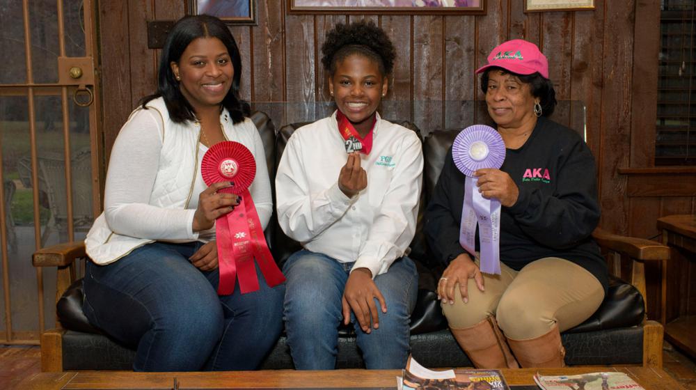 A young female dressed in a white polo and blue jeans smiles and holds up the second-place ribbon around her neck. On the left is an adult woman dressed in a white sheer top with a white vest and blue jeans and is holding a bigger red ribbon. On the right is an older woman wearing a pink AKA hat and a black long-sleeved AKA T-shirt with brown slacks, and she holds up a purple ribbon.