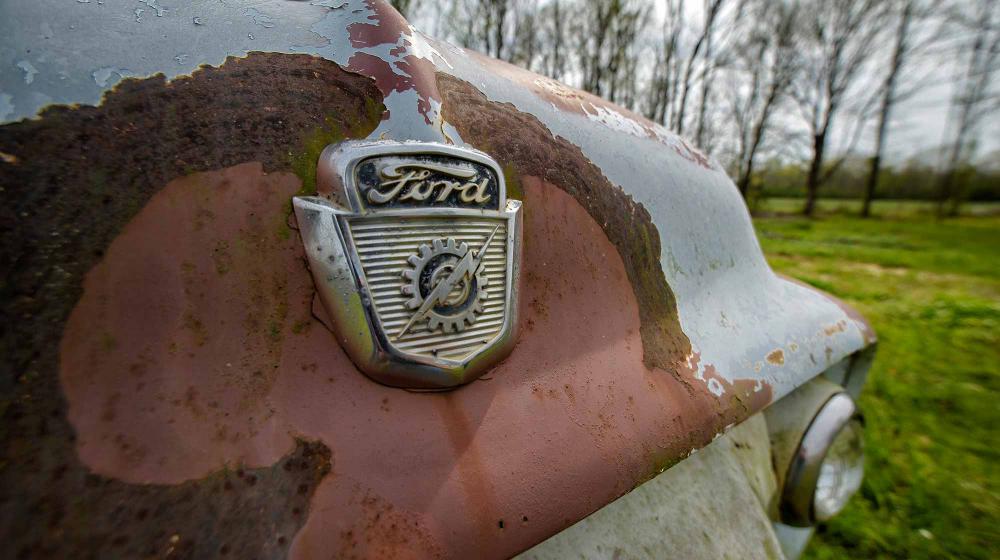 The front of an old Ford truck with the logo centered in an orange-colored rusty area of the light blue truck. 