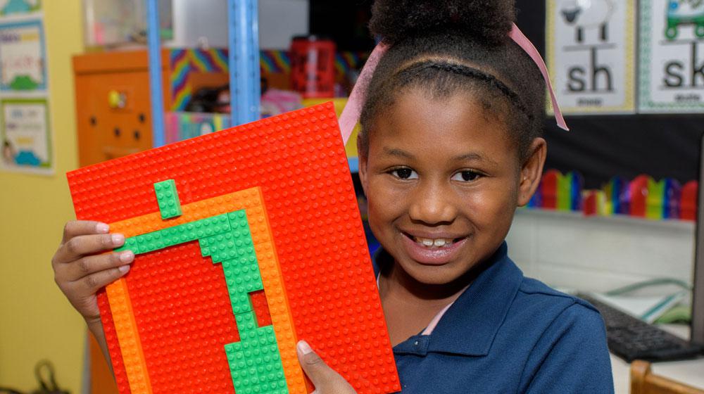 A young girl smiles and holds up a Lego creation.