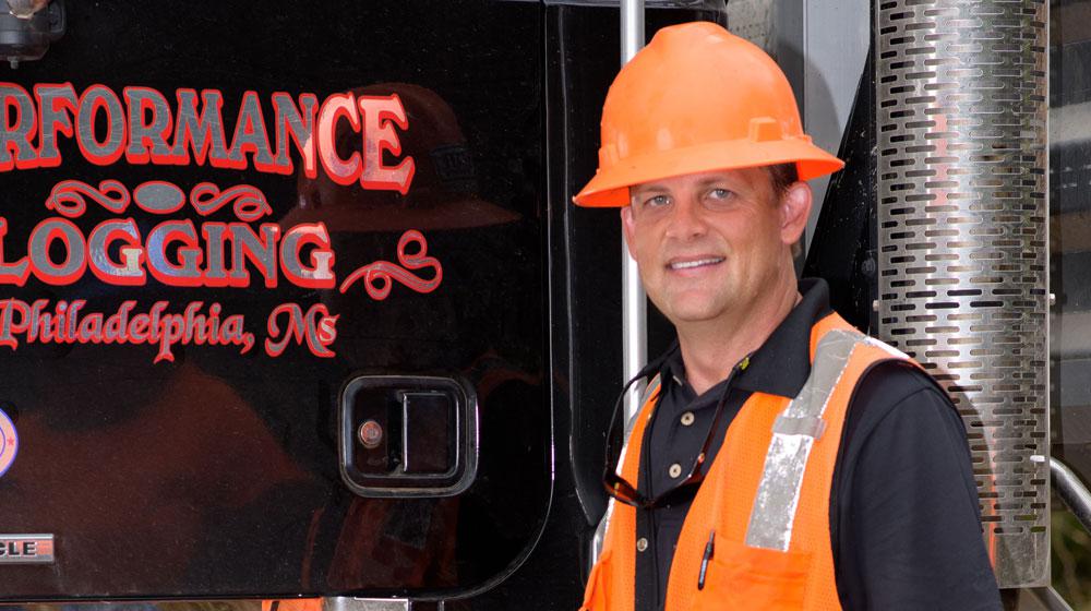 A man wearing a bright orange construction vest and hard hat stands in front of a logging machine.
