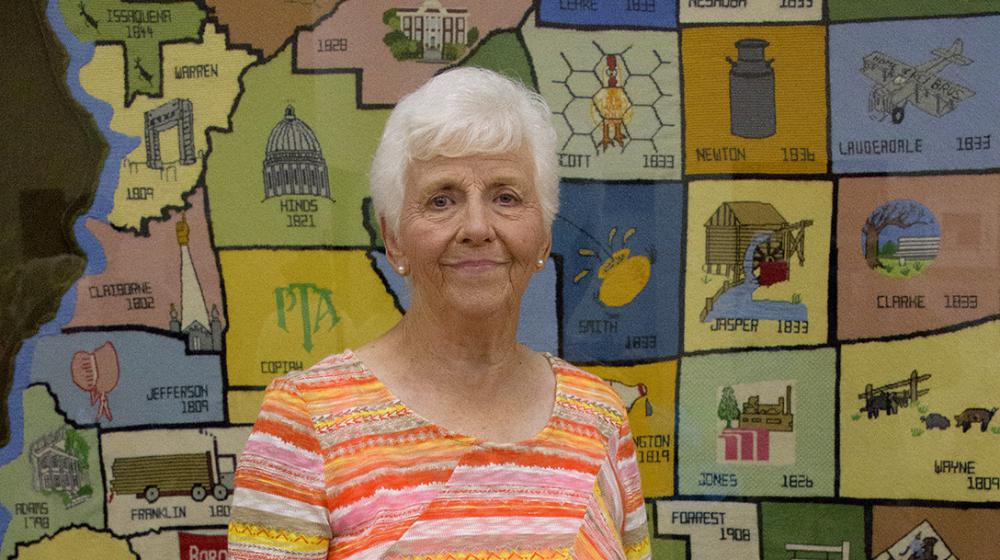An elderly woman wearing an orange striped shirt stands in front of a large, multicolored, needlepoint county map of Mississippi.