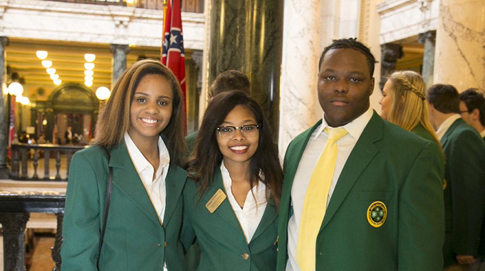 Two young women and one young man wearing green blazers stand on a marble floor.