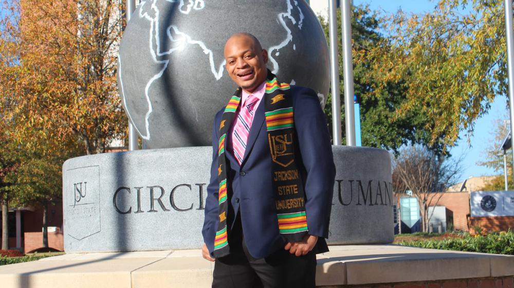 A man wearing a Jackson State University scarf and a navy suit jacket smiles in front of a sculpture on the JSU campus.