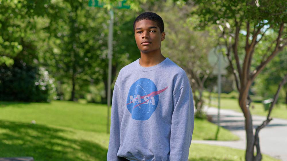 A young boy wearing a NASA sweatshirt stands on a sidewalk holding a camera by his side.