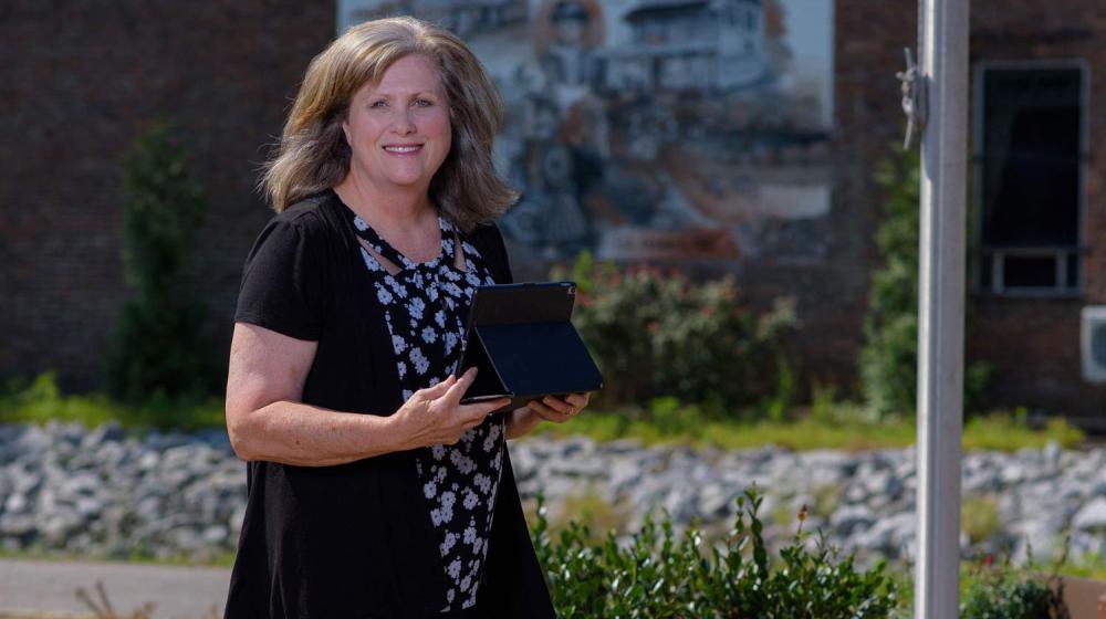 A smiling woman holding a tablet stands outside in front of a flagpole.