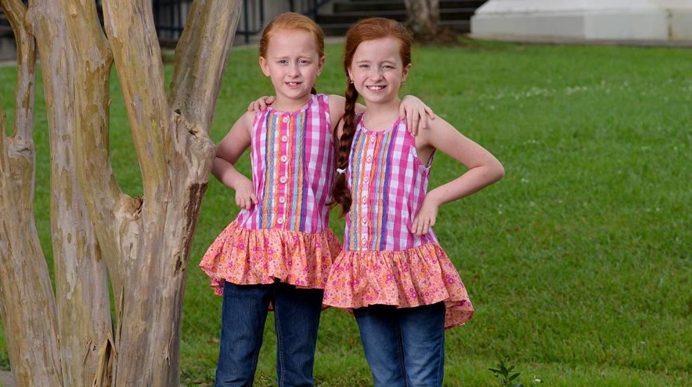 Two young girls with red hair and matching pink shirts stand next to each other with a hand on the hip.