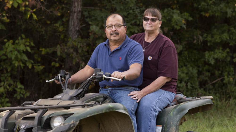 A man wearing a blue shirt and a woman wearing a maroon shirt sitting on a four-wheeler in tall green grass in front of dark green trees.