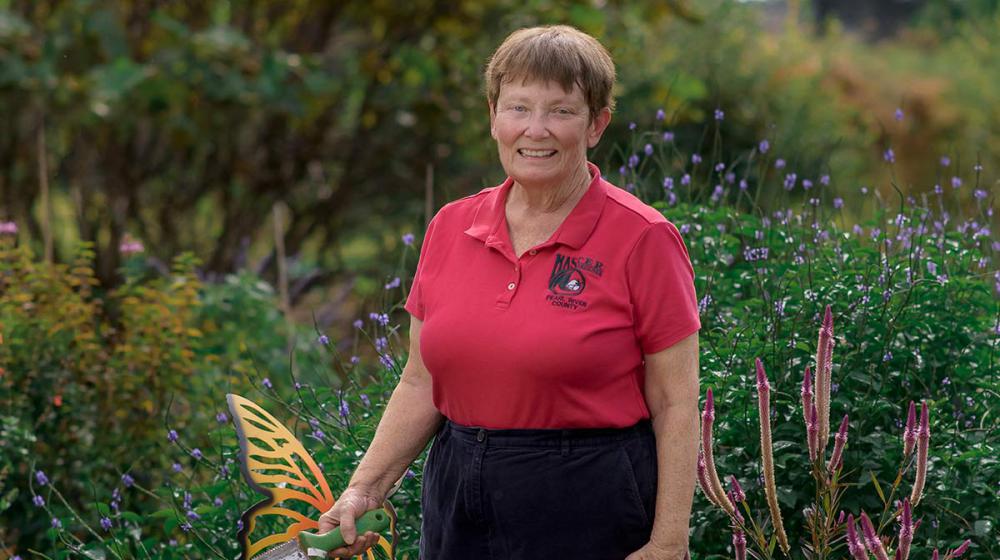 A woman wearing a red collared shirt standing in tall green grass and flowers. She holds a shovel in her right hand, which rests in front of a metal butterfly garden decoration.