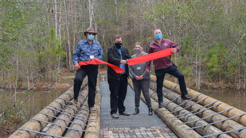 Four people stand behind a recently cut large red ribbon on a new bridge over a body of water.