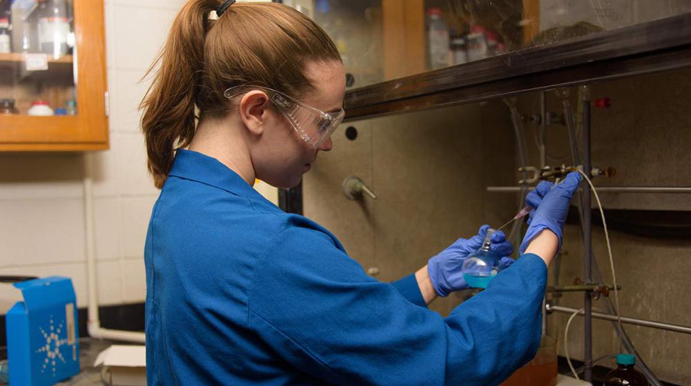 A young woman wearing safety goggles and a blue lab coat working in a lab.
