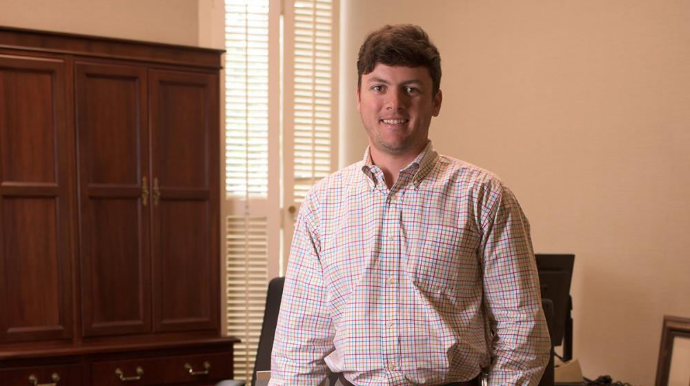 A man wearing a colorful plaid shirt stands in front of a desk.