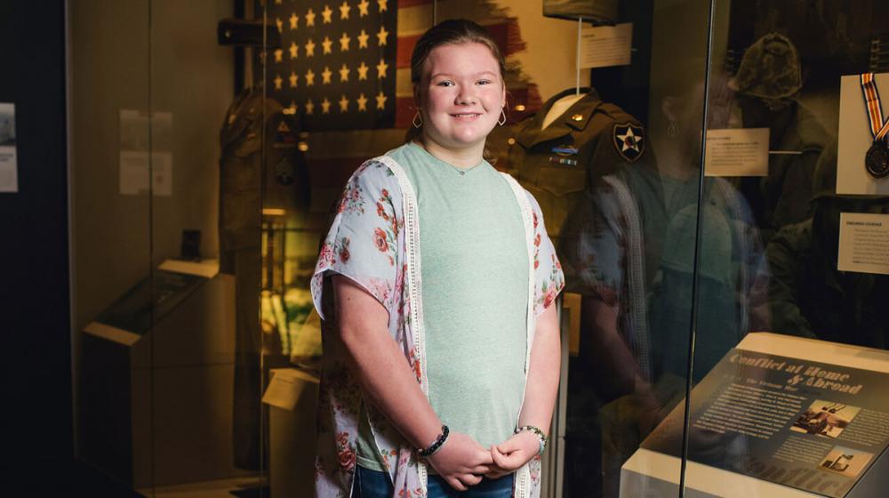A smiling young girl wearing a floral blouse standing in front of a military museum exhibit.
