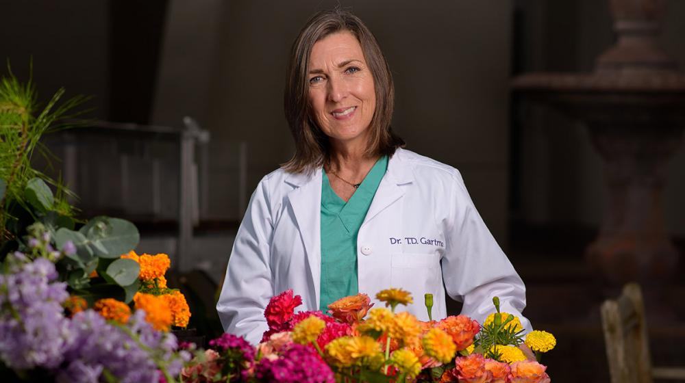 A woman wearing a white coat stands behind floral arrangements and a bucket full of flowers.