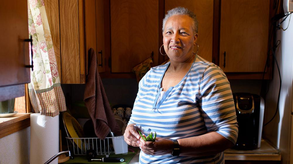 A woman wearing a white and blue striped shirt and holding fresh vegetables stands in front of a kitchen sink.