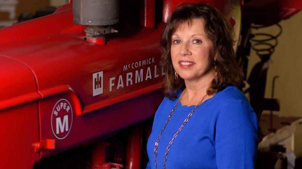 A smiling woman wearing a blue shirt stands next to and rests her arm on a red piece of machinery. 