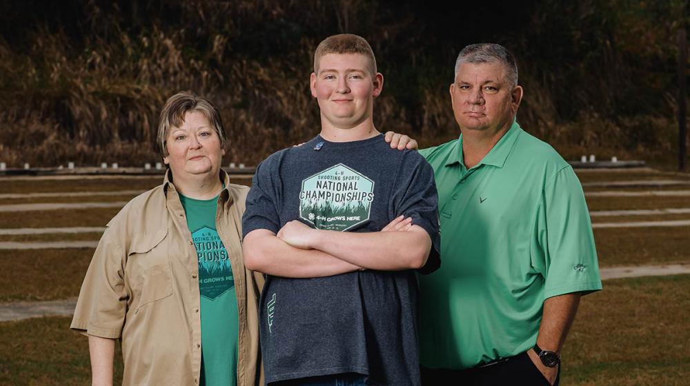 A teenage boy wearing a Shooting Sports National Championships shirt stands between his mother and father as they all smile.