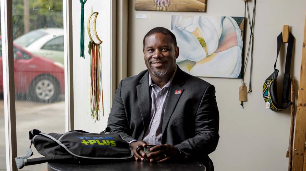 A man wearing a suit sits at a round table with a small mug and a bag that says “Teach Plus.”