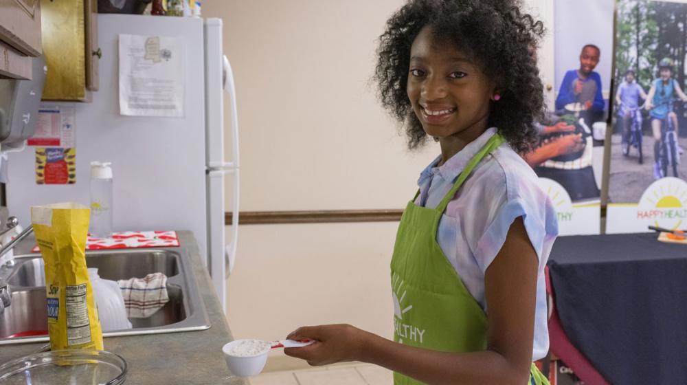 A smiling young girl wearing a green apron holding a measuring cup full of flour.