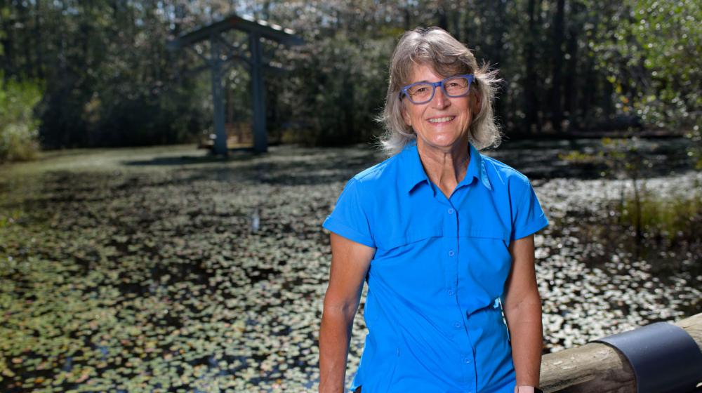 A woman, smiling, wearing a blue polo and sitting on a wooden fence.