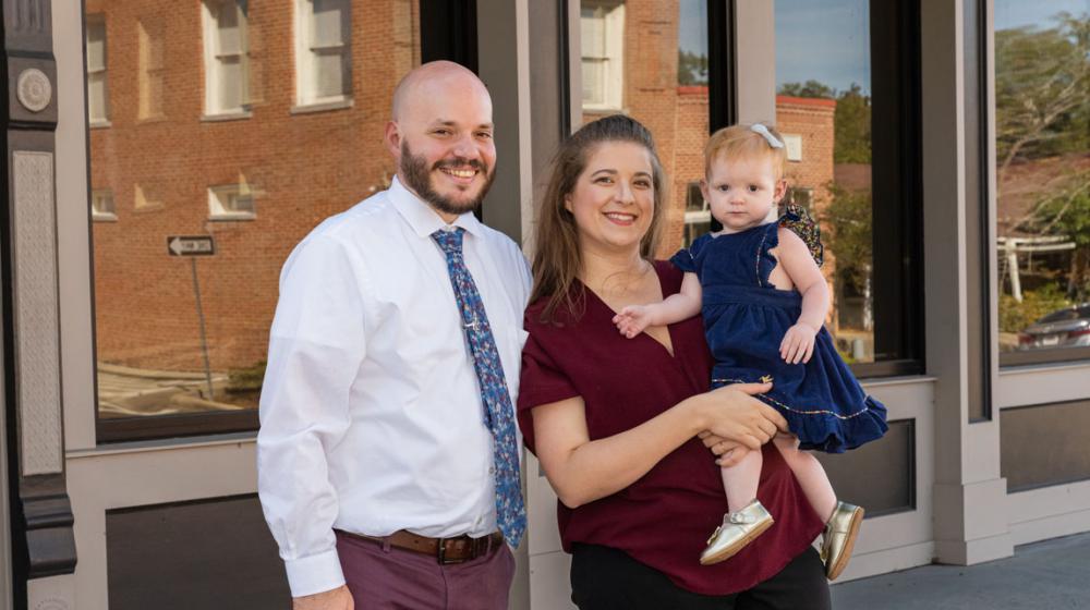 A man, woman, and child standing outside, smiling in front of a building.