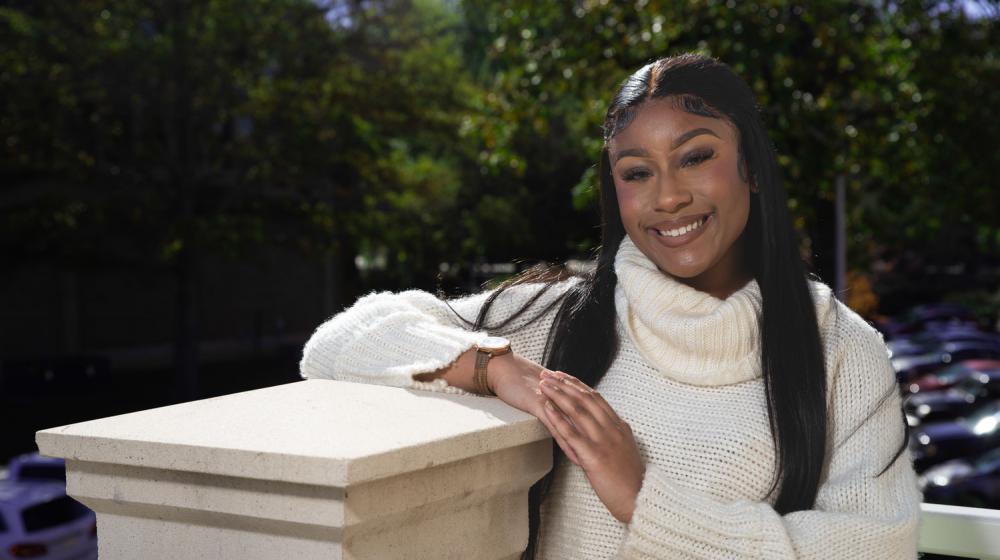 A Black woman wearing a white turtleneck sweater smiling and leaning on a pillar.