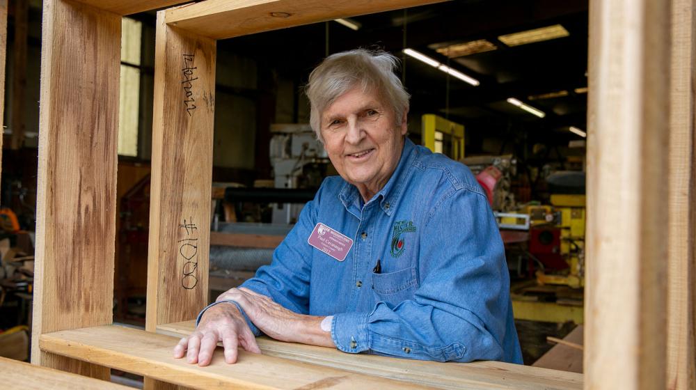 A man wearing a denim shirt and a Master Gardener nametag smiling with a table frame around him listing #1,000 on wood.