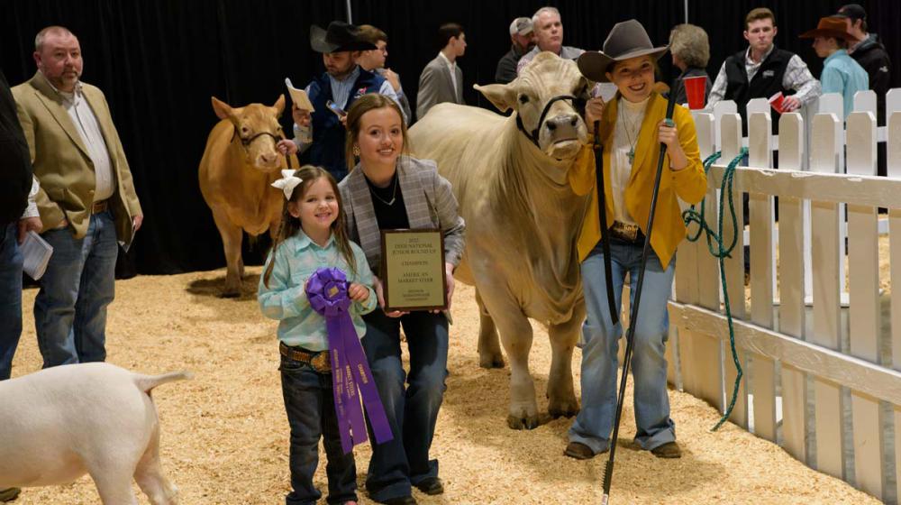 A woman crouches, holding a plaque, with a girl on her left and a boy and steer on her right.