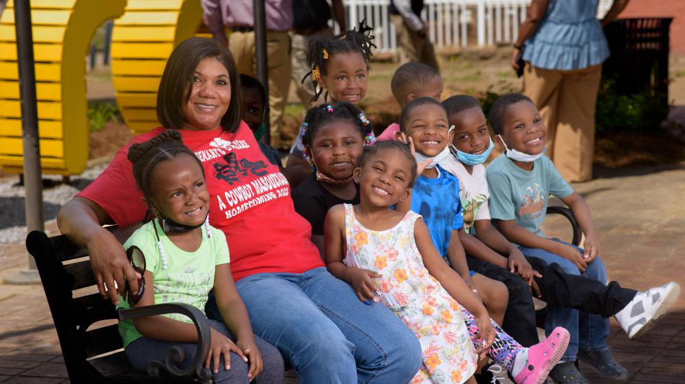 A woman, wearing a red T-shirt listing Rosemary’s Daycare on it, smiles while sitting on a bench with many small children sitting beside her, also smiling.