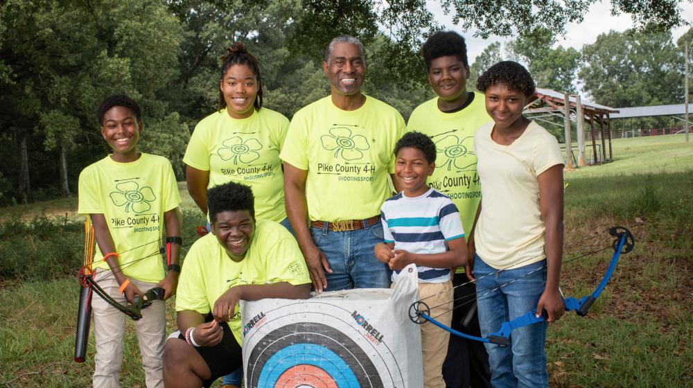 A man standing behind an archery target and surrounded by six children, all smiling.