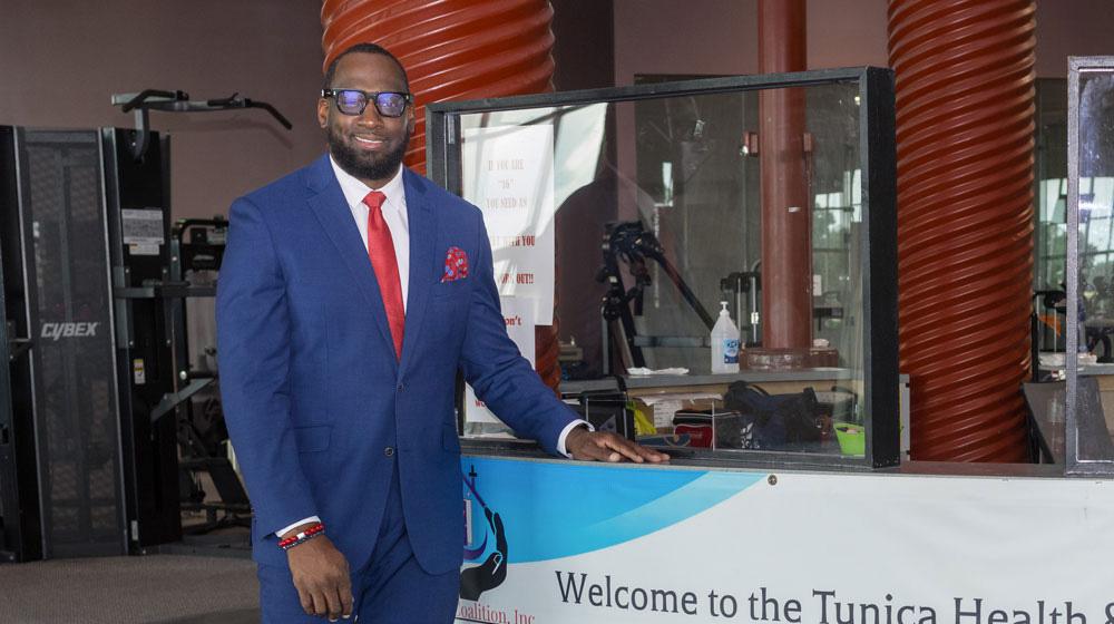 A man wearing a suit stands beside a desk with a banner listing “Welcome to the Tunica Health & Wellness Hub.”