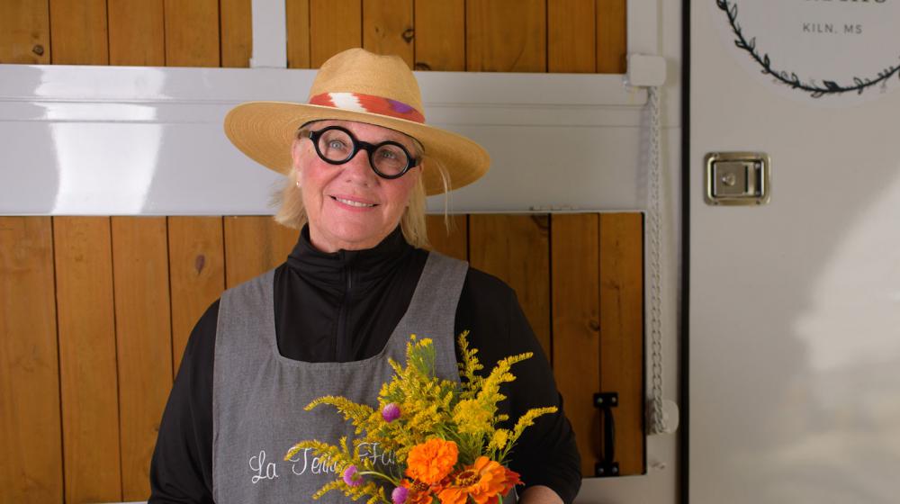 A woman with a straw hat and round-framed glasses holding a bunch of flowers and smiling.