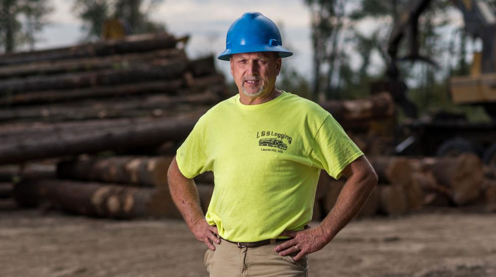 A man, smiling, wearing a hard hat and neon shirt with L&S Logging printed on it.