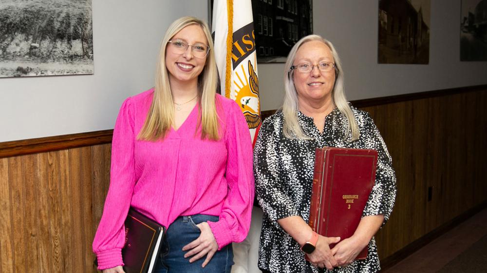 Two women standing in front of a flag, smiling.