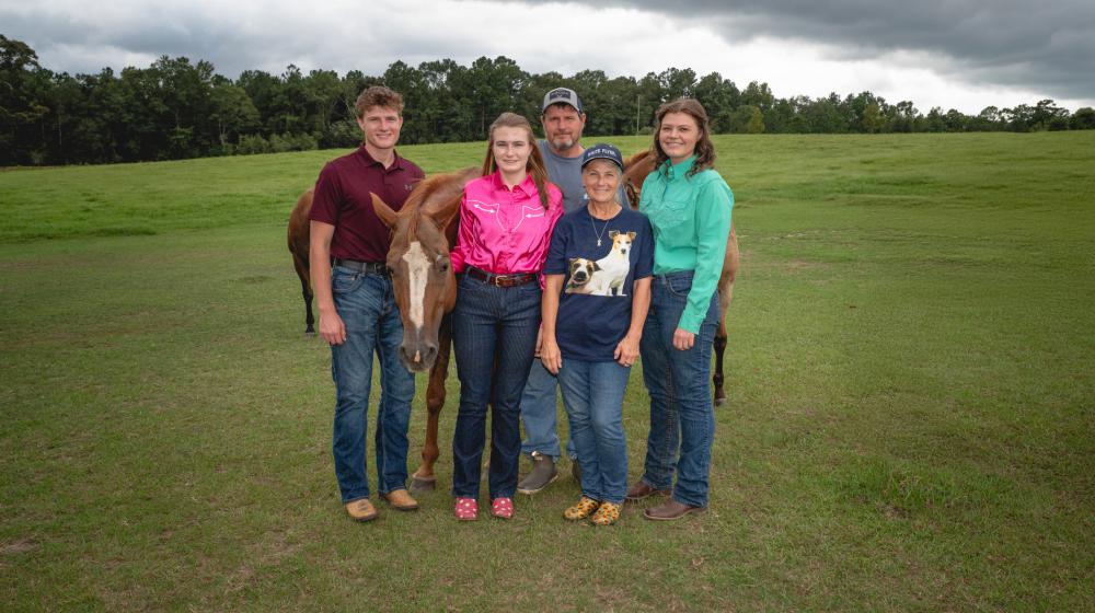 Five people smiling with three horses, all standing on a grassy plain